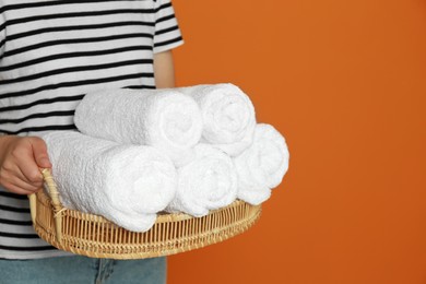 Photo of Woman holding wicker tray with rolled soft terry towels on orange background, closeup. Space for text