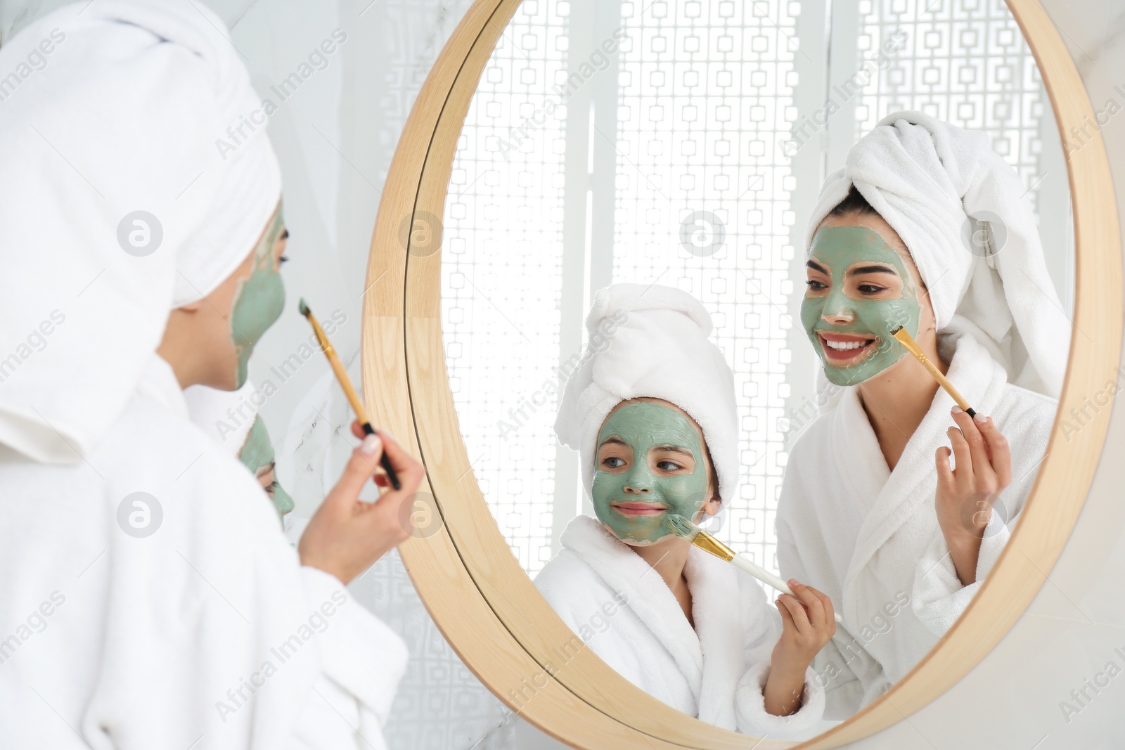 Photo of Young mother and her daughter with facial masks near mirror in bathroom