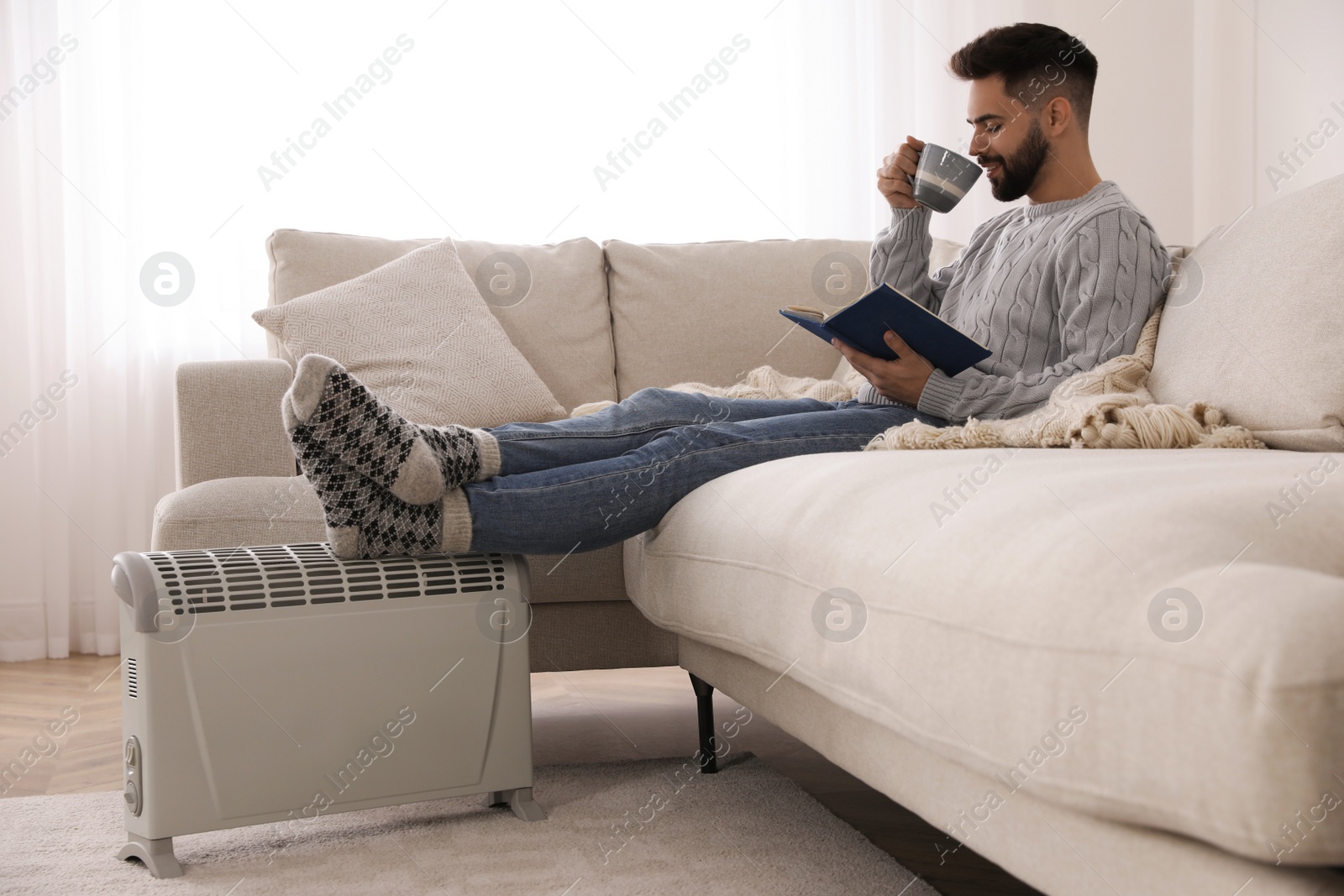 Photo of Young man with book and hot drink warming feet on electric heater at home