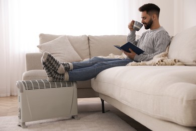 Young man with book and hot drink warming feet on electric heater at home