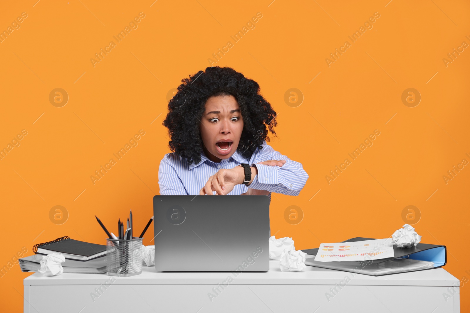Photo of Stressful deadline. Scared woman checking time on wristwatch at white table against orange background