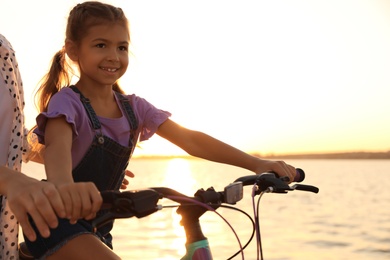 Happy mother teaching her daughter to ride bicycle near river at sunset