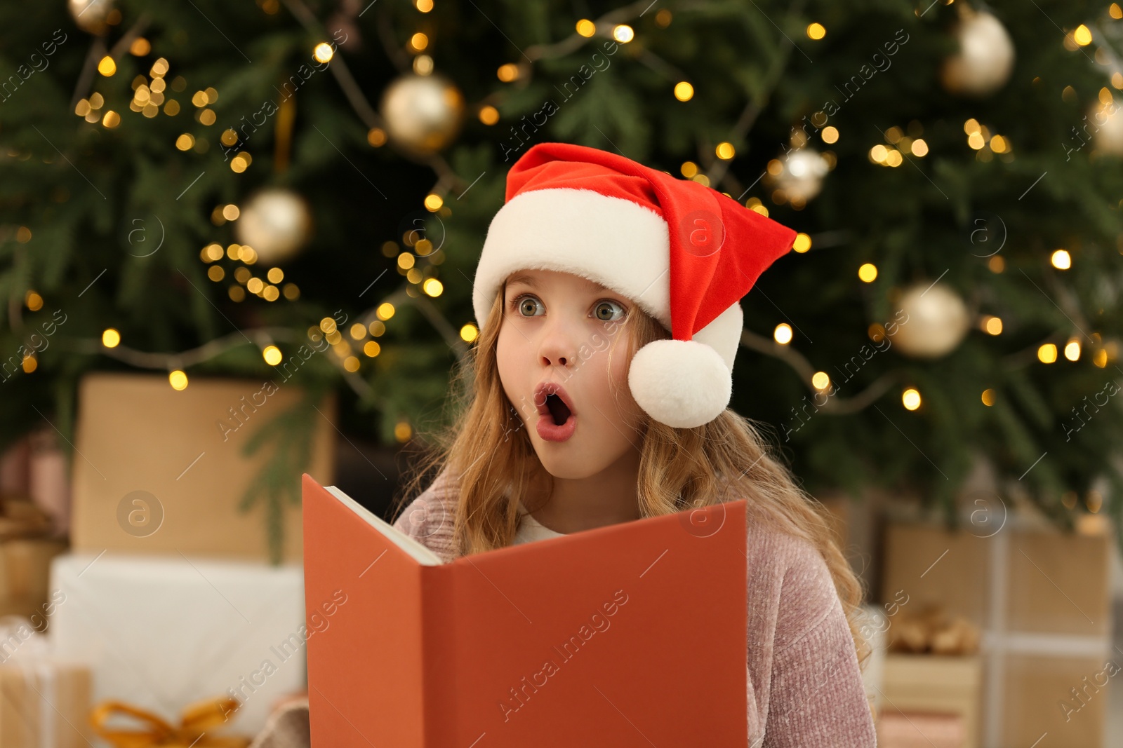 Photo of Emotional little girl in Santa hat with book near tree and gifts at home. Christmas atmosphere