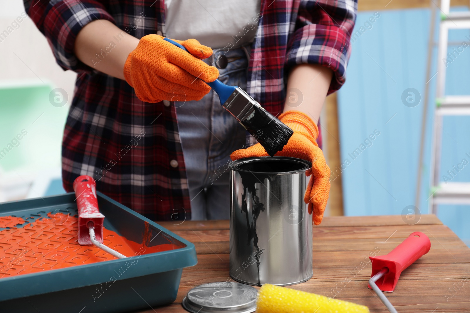Photo of Woman dipping brush into can of black paint at wooden table indoors, closeup