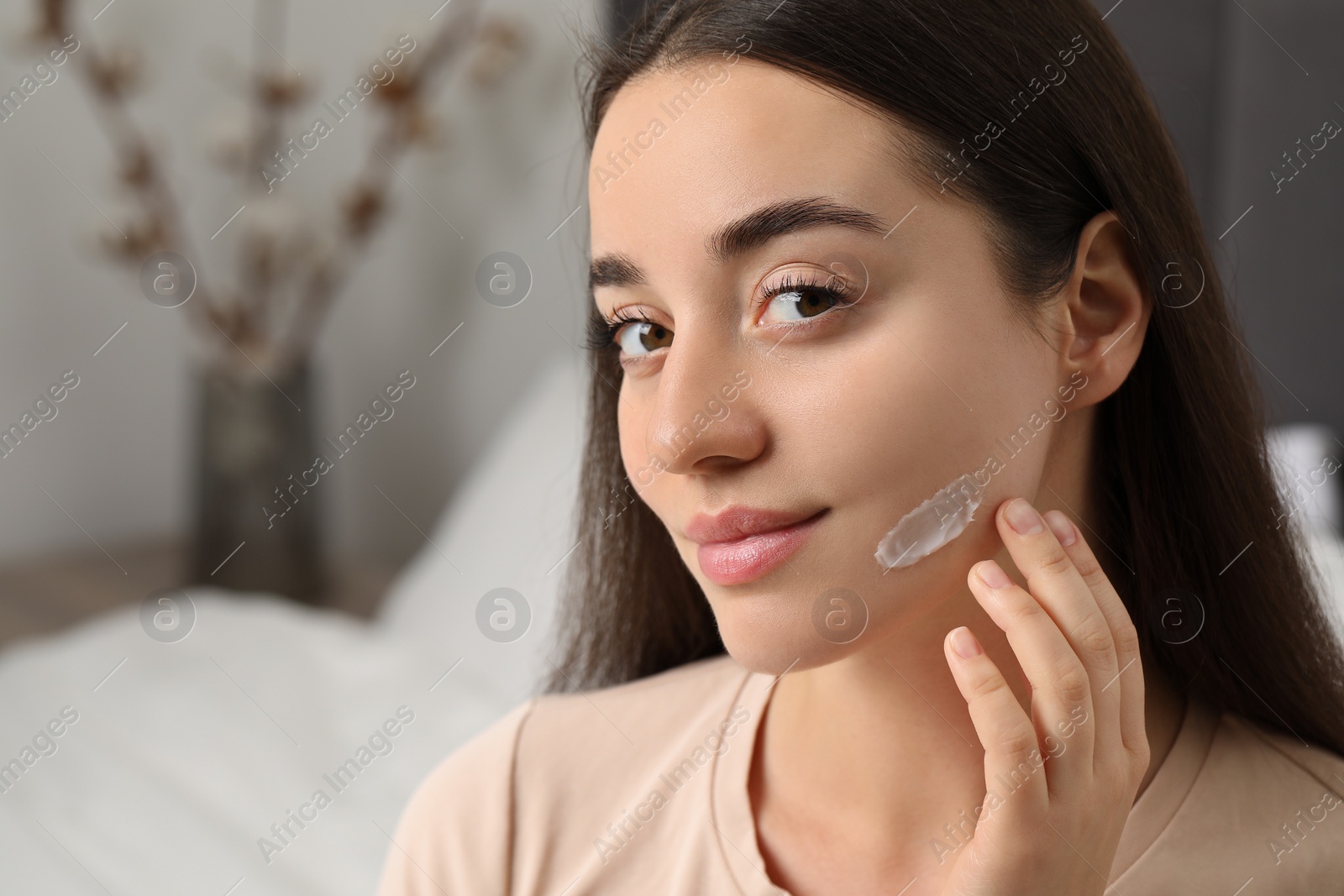 Photo of Young woman with dry skin applying cream onto her face indoors. Space for text