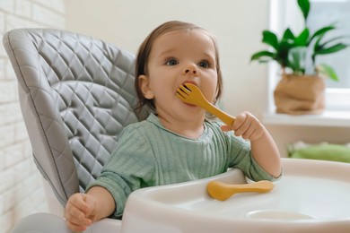 Photo of Cute little baby nibbling fork in high chair indoors