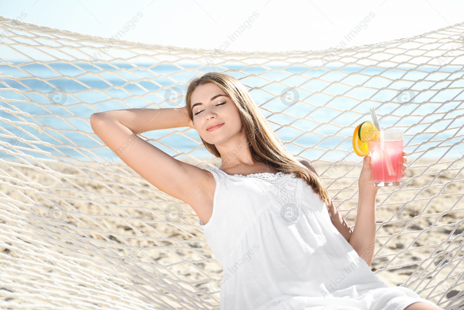 Photo of Young woman with refreshing cocktail relaxing in hammock on beach