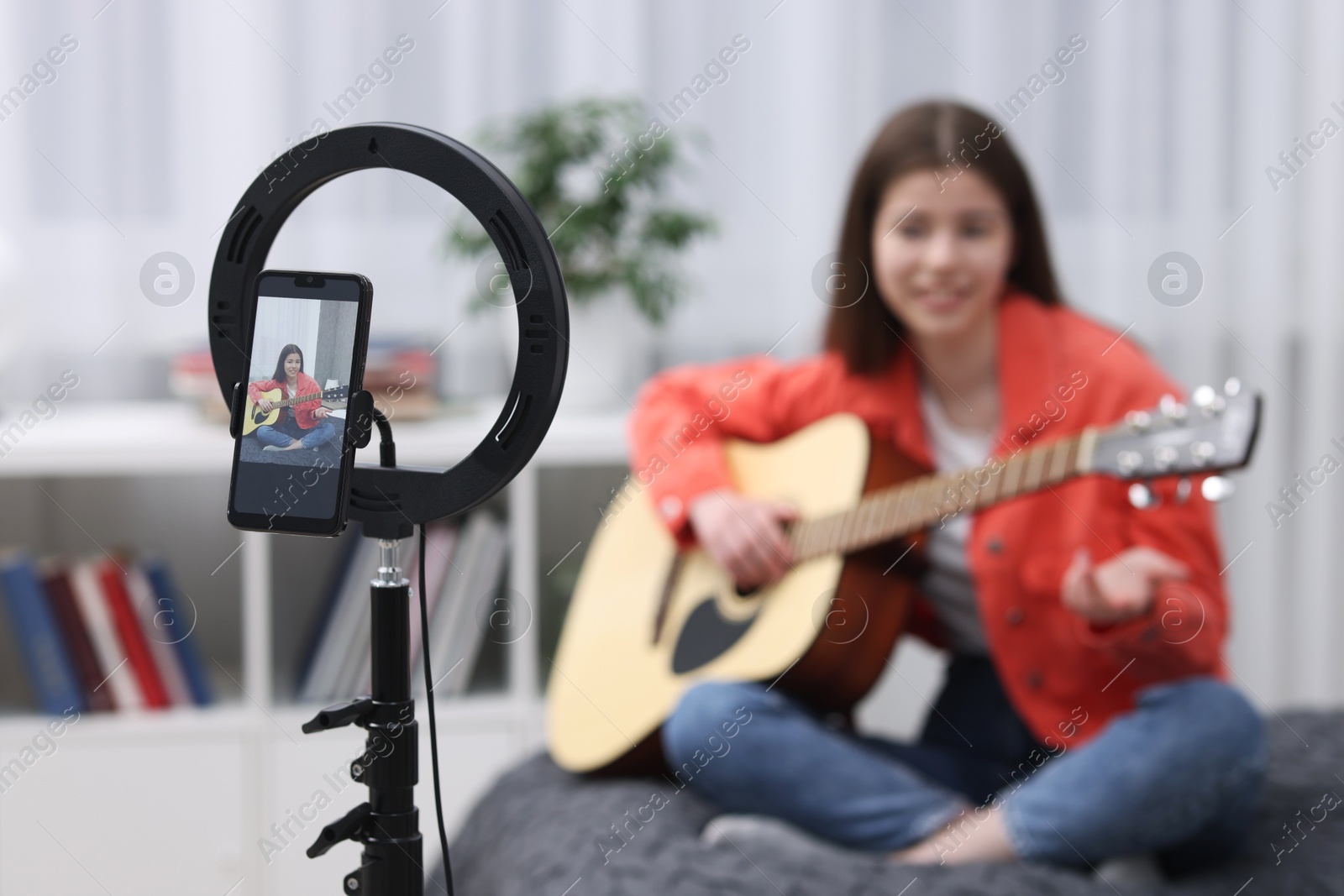 Photo of Teenage blogger playing guitar while streaming at home, focus on smartphone