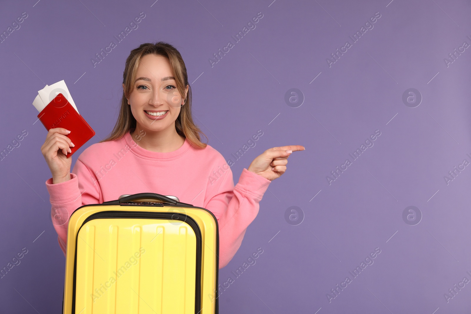 Photo of Happy young woman with passport, ticket and suitcase pointing at something on purple background, space for text