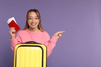 Happy young woman with passport, ticket and suitcase pointing at something on purple background, space for text