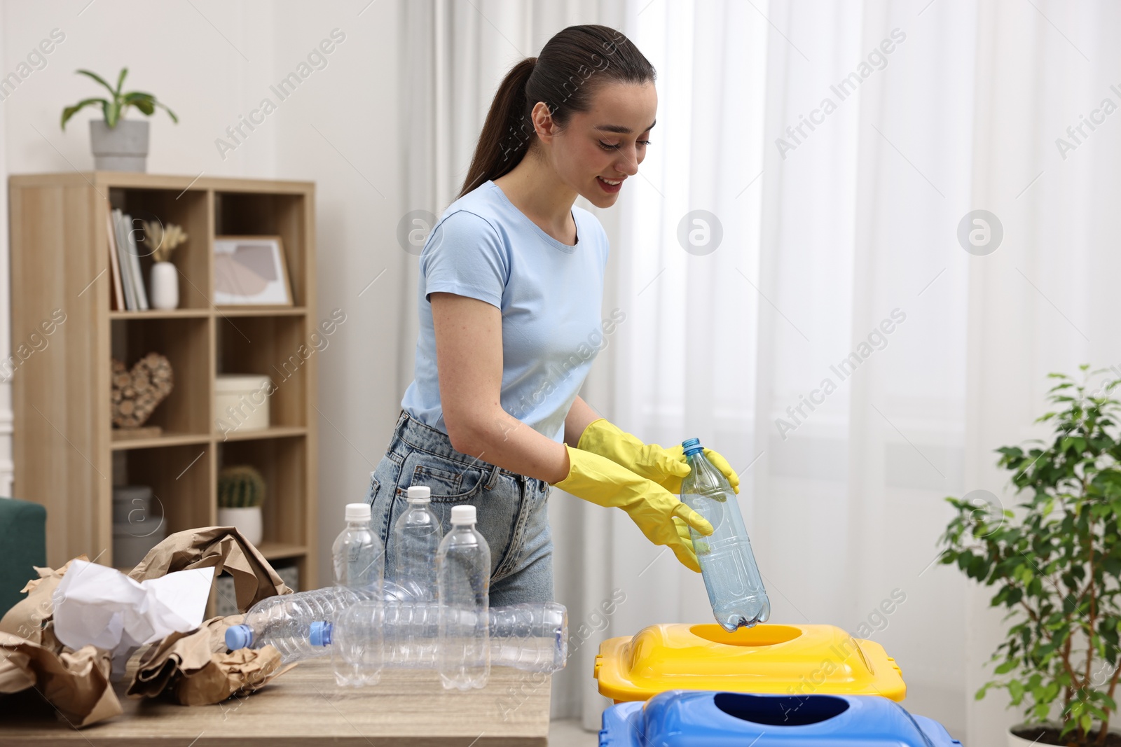 Photo of Garbage sorting. Smiling woman throwing plastic bottle into trash bin in room