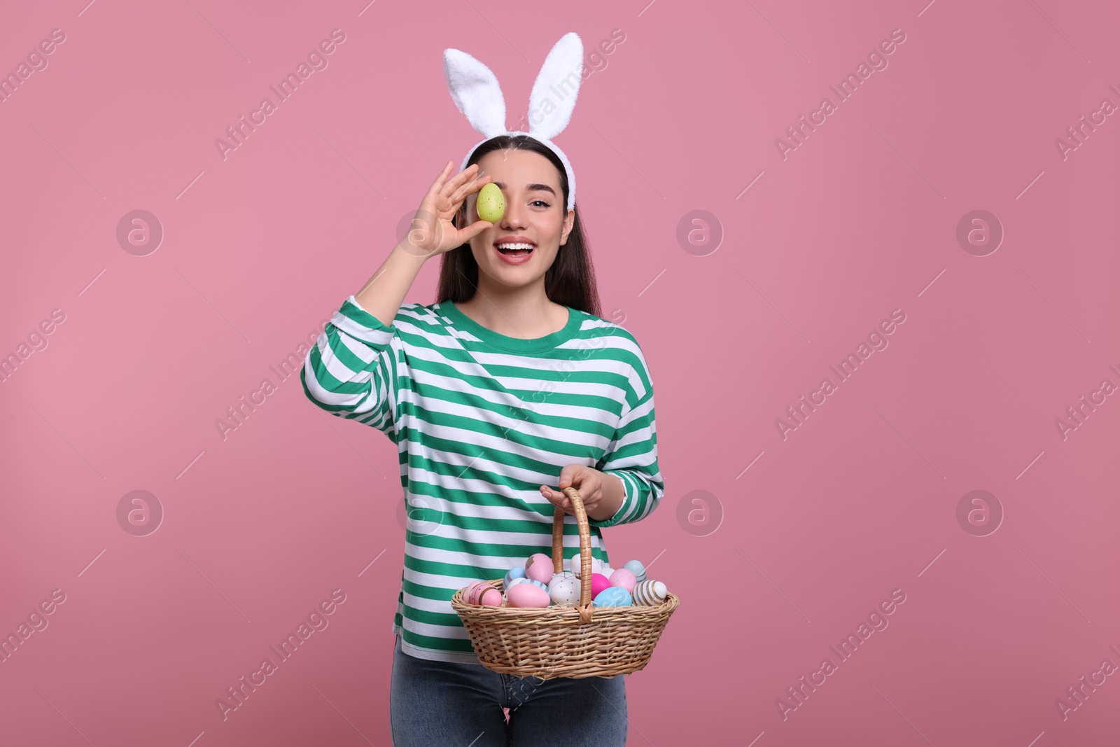 Photo of Happy woman in bunny ears headband holding wicker basket of painted Easter eggs on pink background