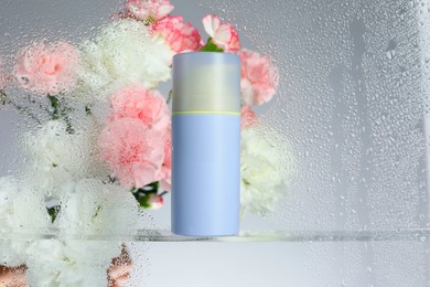 Photo of Bottle with moisturizing cream and beautiful flowers on light background, view through wet glass. Space for text