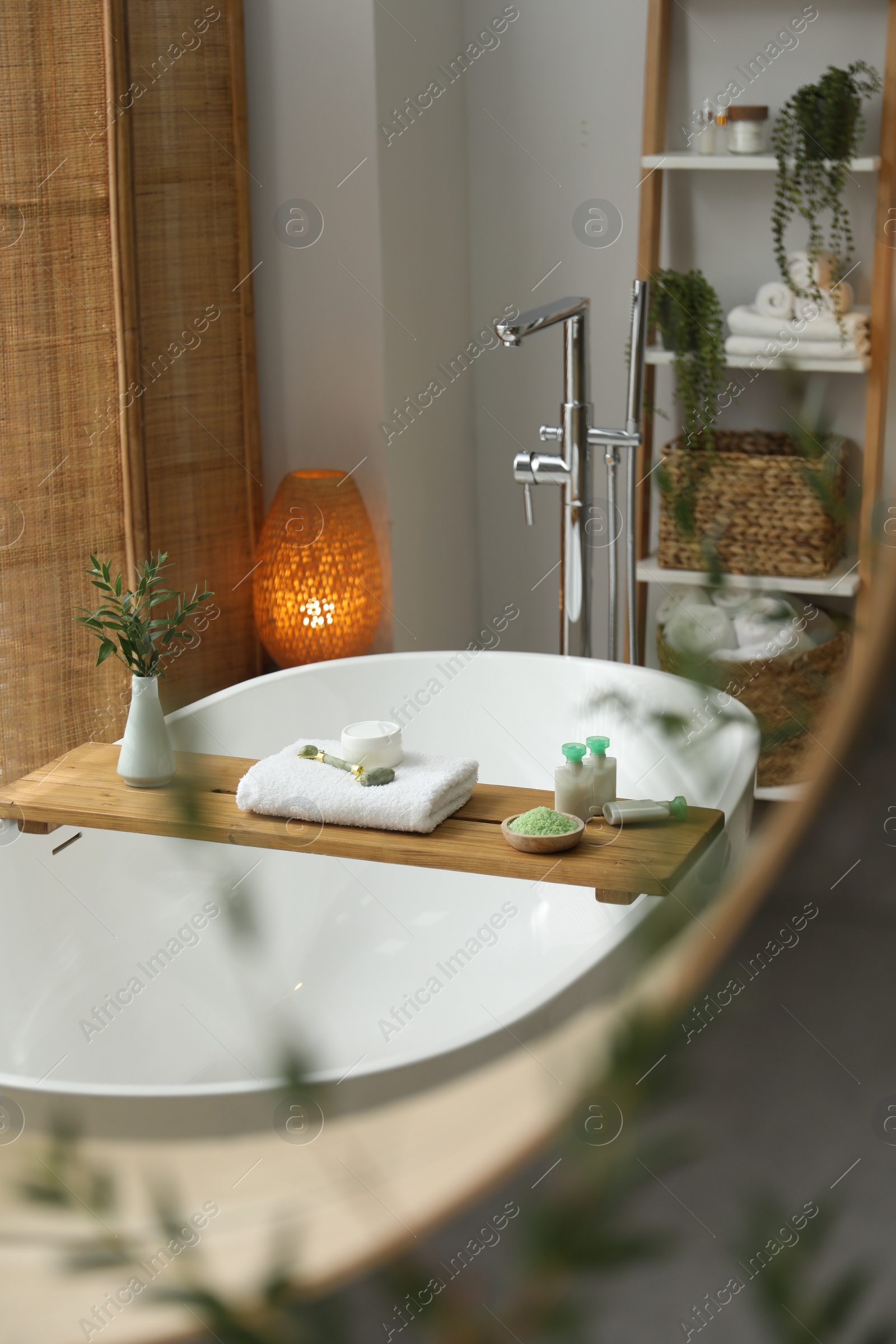 Photo of Wooden tray with spa products and green branches on bath tub in bathroom, reflection in mirror