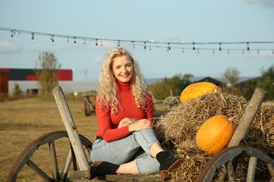 Beautiful woman sitting on wooden cart with pumpkins and hay in field. Autumn season