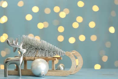 Photo of Wooden sleigh with small fir tree near decorative reindeer and Christmas ornament on light blue table against blurred lights. Space for text