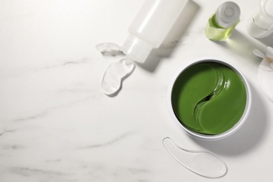 Jar of under eye patches with spoon and cosmetic products on white marble table, flat lay. Space for text