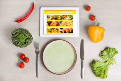 Photo of Modern tablet with open page for online food ordering, scattered vegetables, plate and cutlery on white wooden table, flat lay. Concept of delivery service