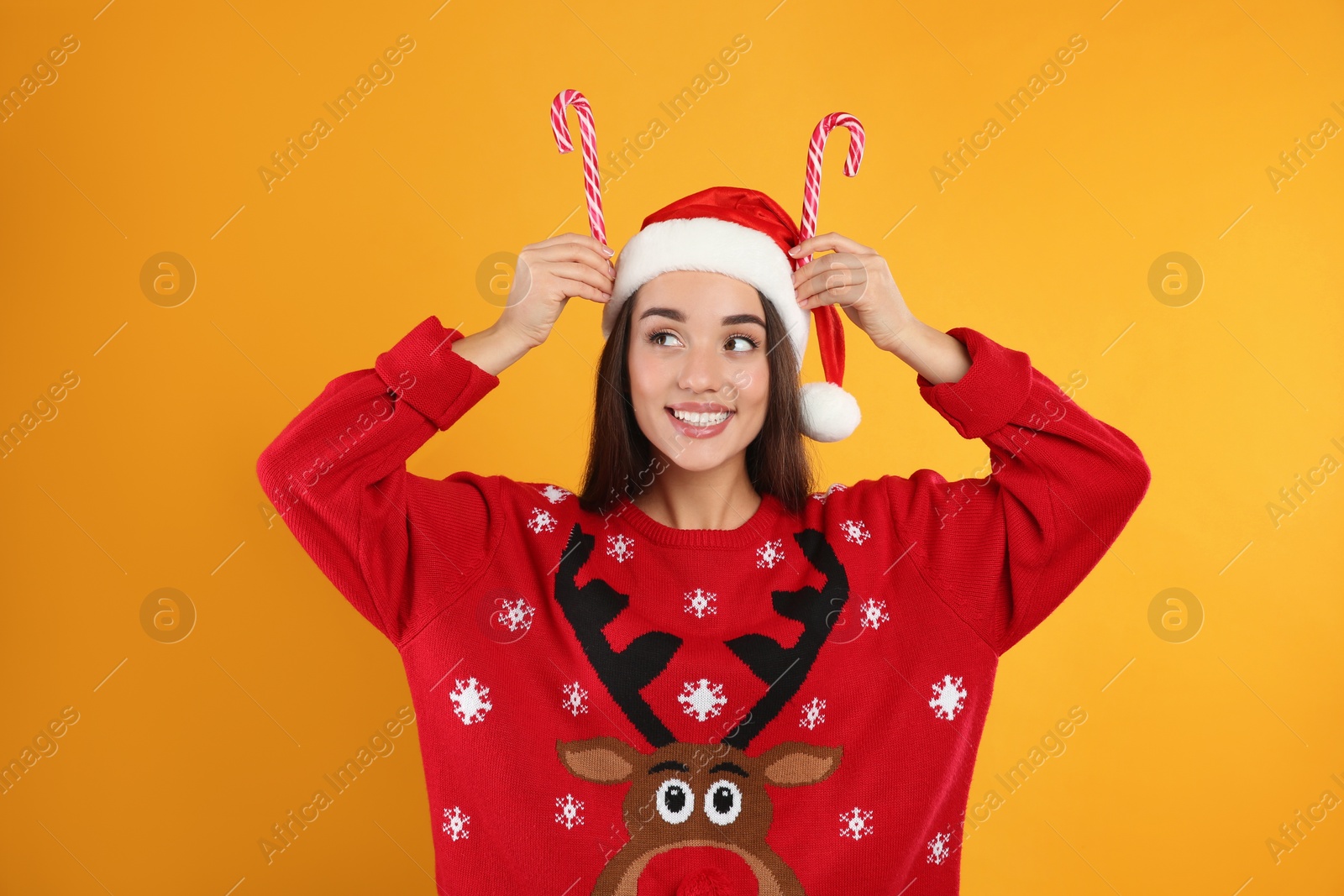 Photo of Young woman in Christmas sweater and Santa hat holding candy canes on yellow background