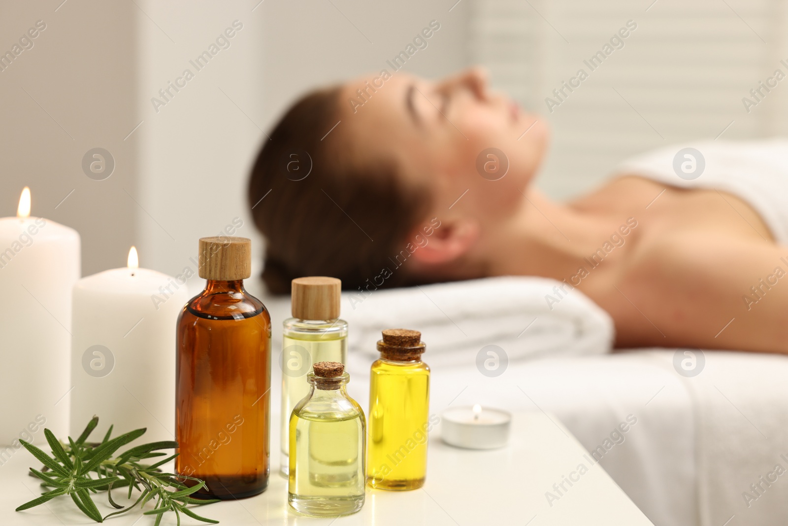 Photo of Aromatherapy. Woman relaxing on massage couch in spa salon, focus on bottles of essential oils, burning candles and rosemary