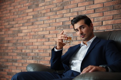 Young man with glass of whiskey near brick wall indoors. Space for text