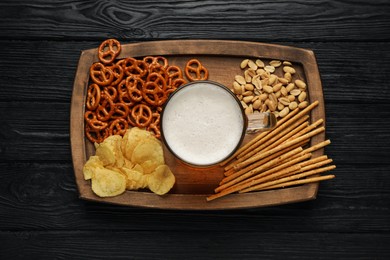Glass of beer served with delicious pretzel crackers and other snacks on black wooden table, top view