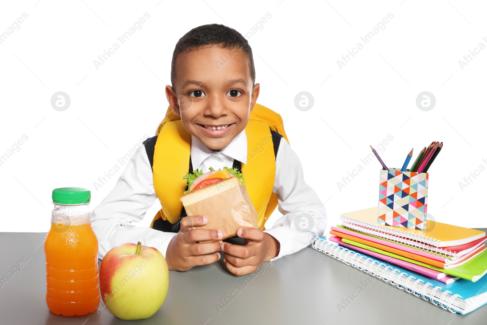 Photo of African-American schoolboy with healthy food and backpack sitting at table on white background