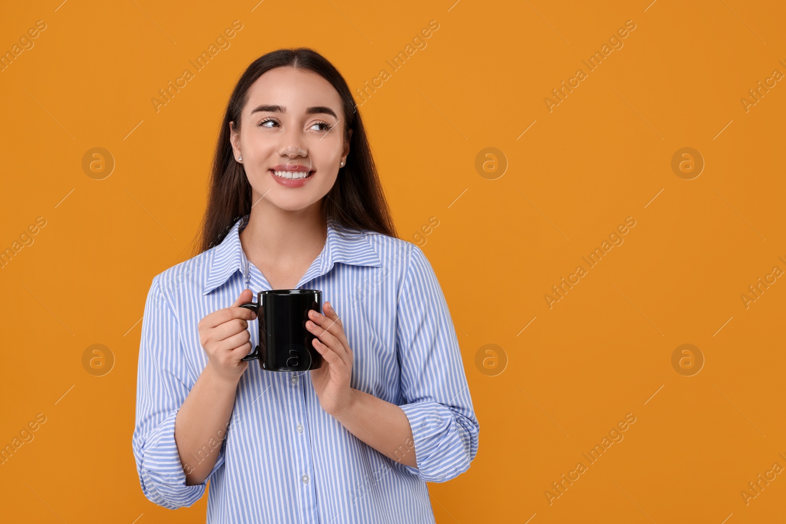 Photo of Happy young woman holding black ceramic mug on orange background, space for text