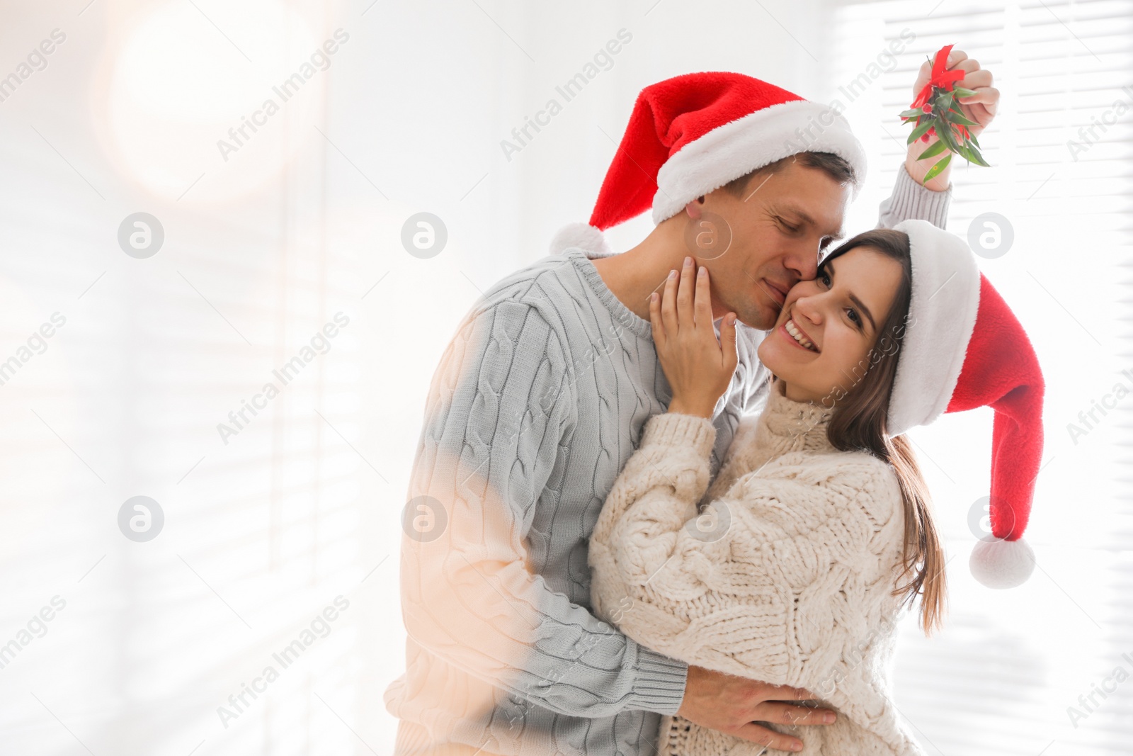 Photo of Happy man kissing his girlfriend under mistletoe bunch at home, bokeh effect. Space for text