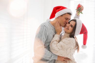 Photo of Happy man kissing his girlfriend under mistletoe bunch at home, bokeh effect. Space for text