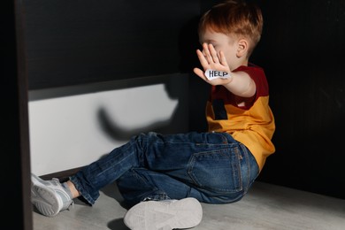 Photo of Scared little boy holding piece of paper with word Help on floor at home. Domestic violence concept