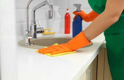 Photo of Woman in protective gloves cleaning kitchen sink with rag, closeup