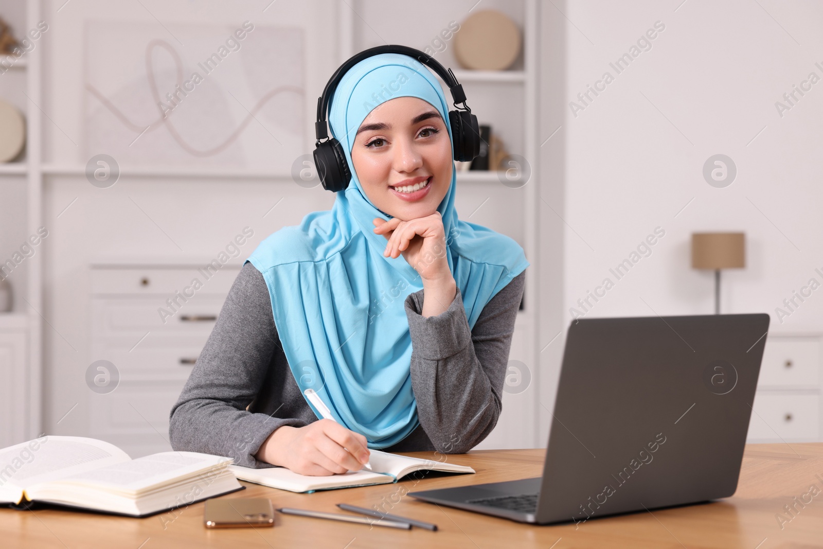 Photo of Muslim woman in headphones writing notes near laptop at wooden table in room