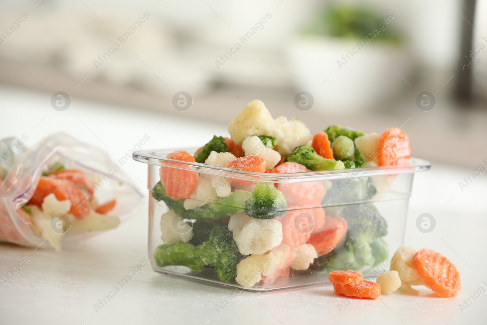 Photo of Mix of different frozen vegetables in plastic container on white textured countertop in kitchen, closeup