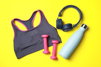 Photo of Stylish sports bra, dumbbells, thermo bottle and headphones on yellow background, flat lay