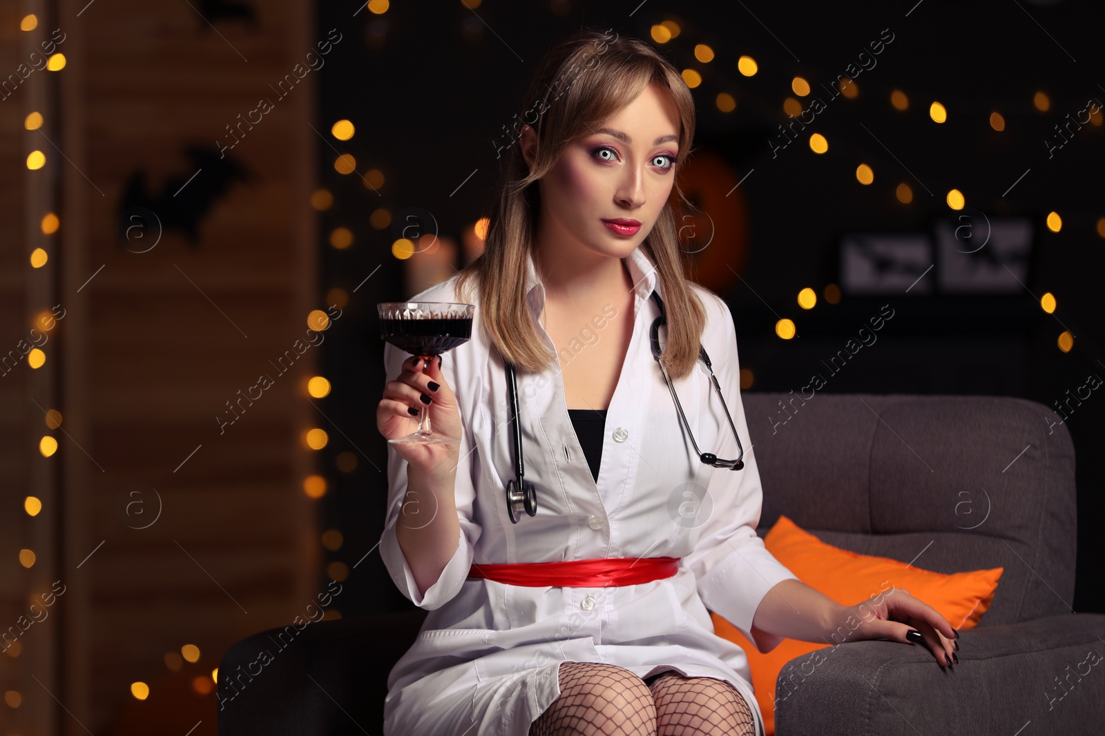 Photo of Woman in scary nurse costume with glass of wine against blurred lights indoors. Halloween celebration