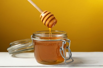 Pouring honey from dipper into jar at white wooden table against golden background