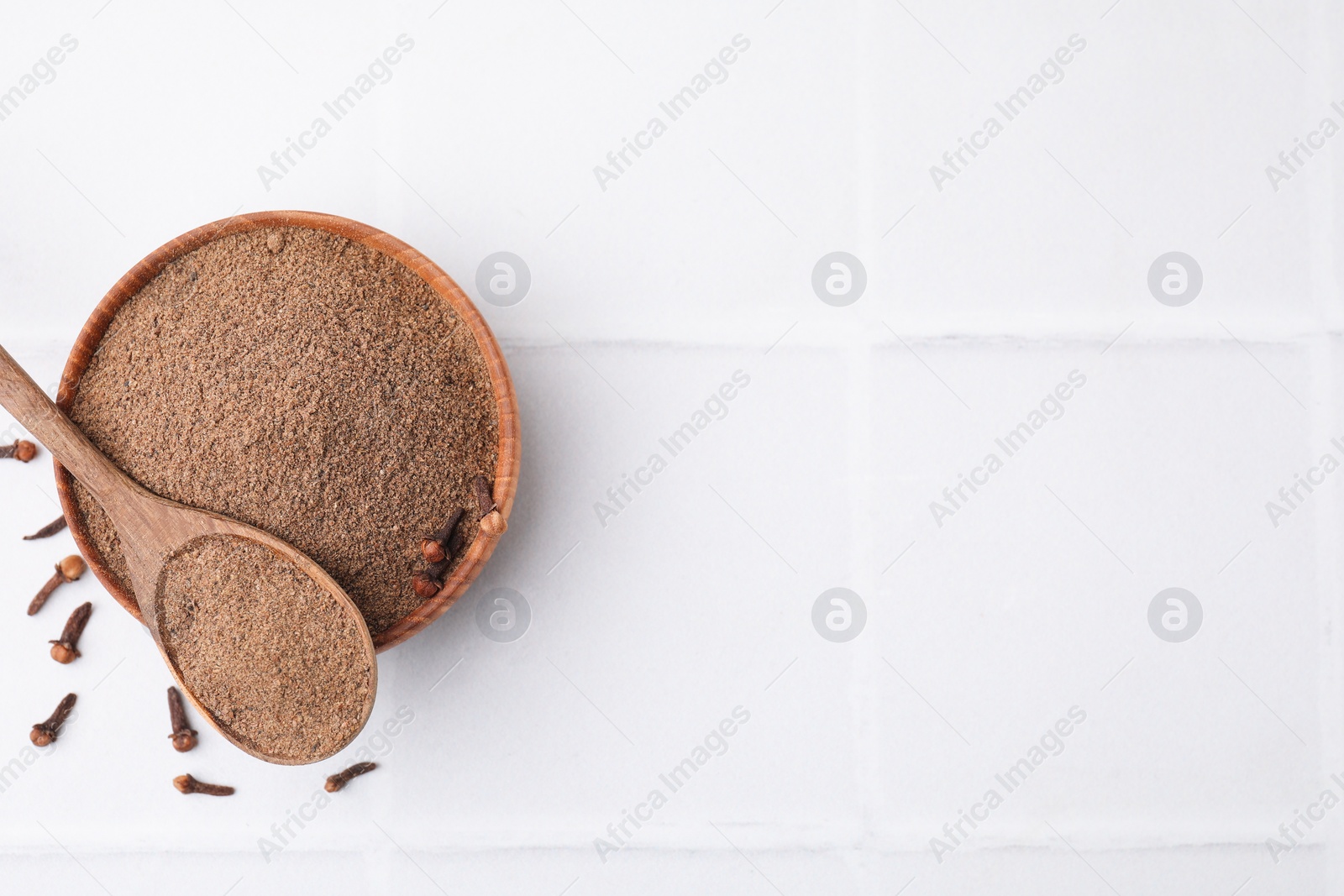 Photo of Aromatic clove powder in bowl, dried buds and spoon on white tiled table, top view. Space for text