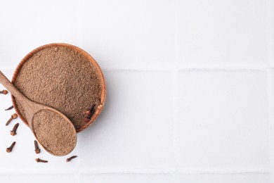 Photo of Aromatic clove powder in bowl, dried buds and spoon on white tiled table, top view. Space for text