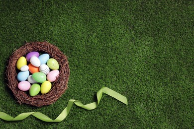 Photo of Nest with many colorful Easter eggs and ribbon on green grass, flat lay. Space for text