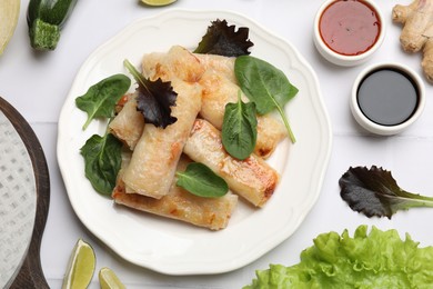 Photo of Plate with tasty fried spring rolls, spinach, sauces and other products on white tiled table, flat lay