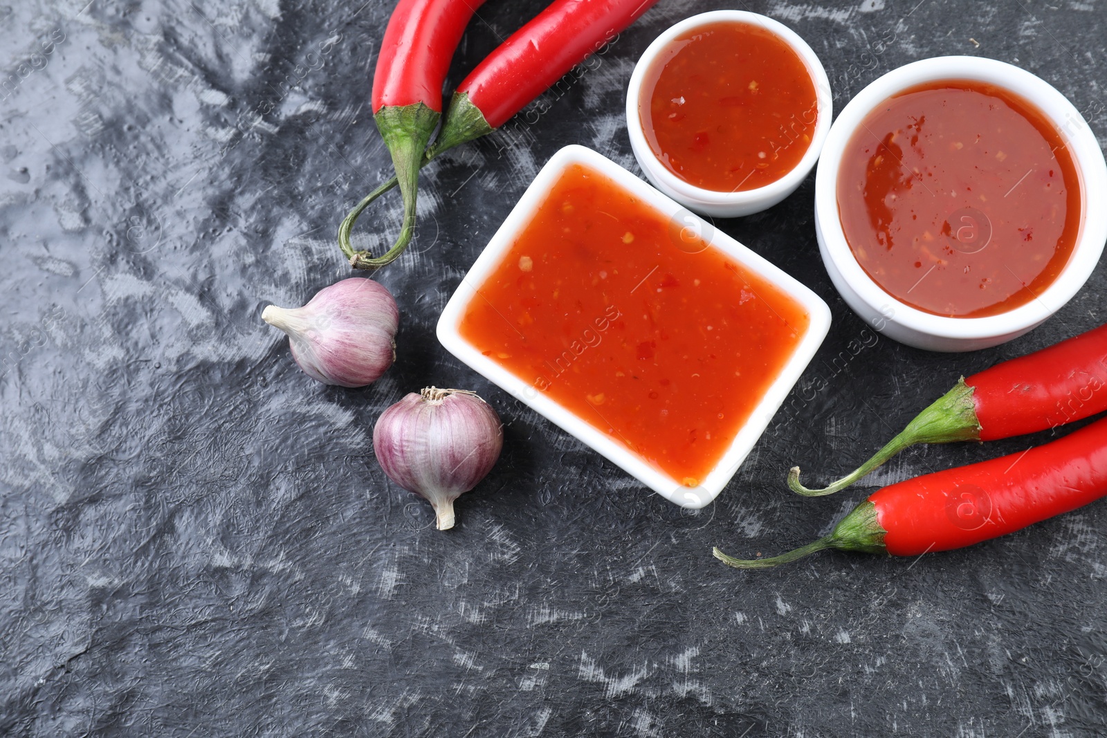 Photo of Spicy chili sauce, peppers and garlic on black textured table, flat lay. Space for text