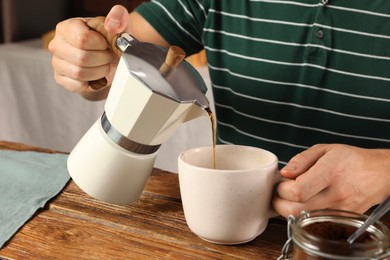 Man pouring aromatic coffee from moka pot into cup at wooden table indoors, closeup