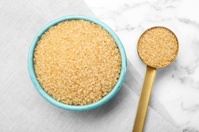 Brown sugar in bowl and scoop on white marble table, top view