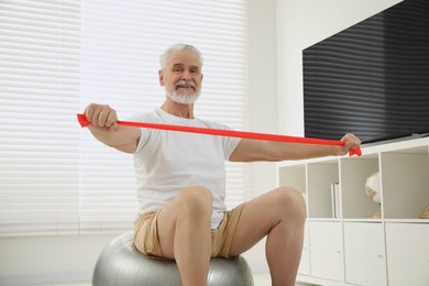 Photo of Senior man doing exercise with elastic resistance band on fitness ball at home