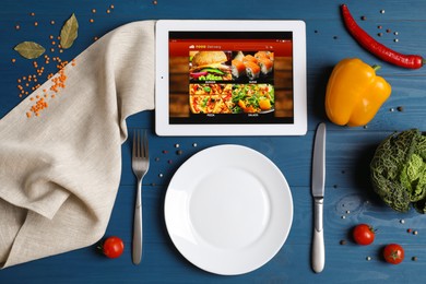 Modern tablet with open page for online food ordering, scattered vegetables, spices, plate and cutlery on blue wooden table, flat lay. Concept of delivery service