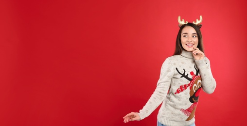 Young woman in Christmas sweater and deer headband on red background, space for text