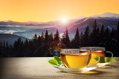 Image of Cups of hot tea on wooden table and beautiful winter landscape on background