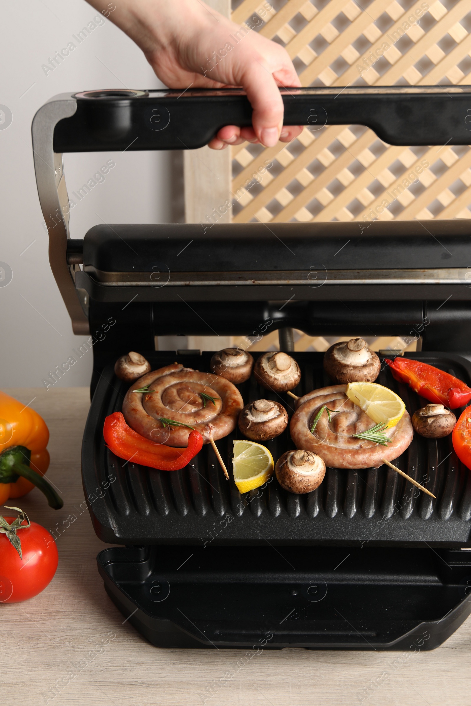 Photo of Woman cooking homemade sausages with bell peppers and mushrooms on electric grill at wooden table, closeup