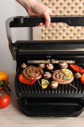 Photo of Woman cooking homemade sausages with bell peppers and mushrooms on electric grill at wooden table, closeup